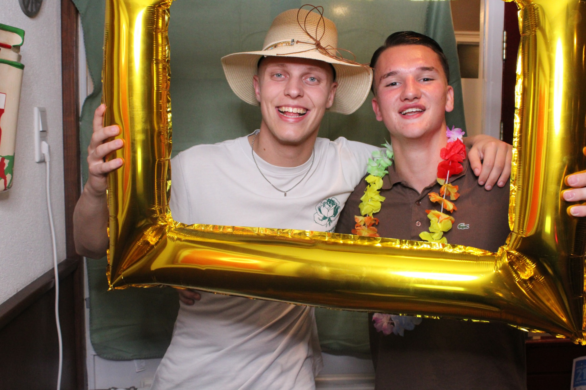 Twee feestgangers met hoed en lei in een gouden ballonnenframe bij de photobooth – verhuur door Uitjes2go in Ede